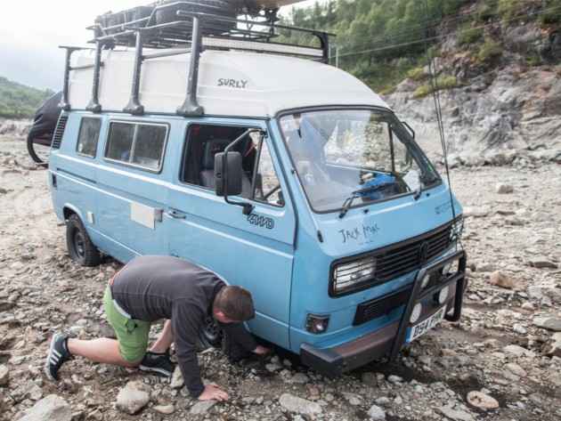 Person bending down next to a flat right front tire of a light blue VW van in a rocky environment