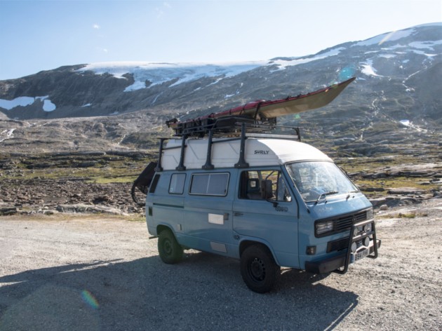 A light blue VW van with kayak on the roof rack drive on a gravel road in the mountains