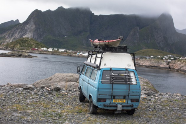 Rear view of a light VW van with a kayak on top, facing a river in the mountains