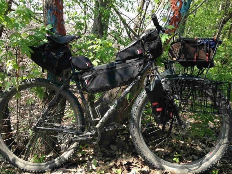 Right side view of a dirty, black Surly Karate Monkey bike loaded with gear, parked against a tree in the woods