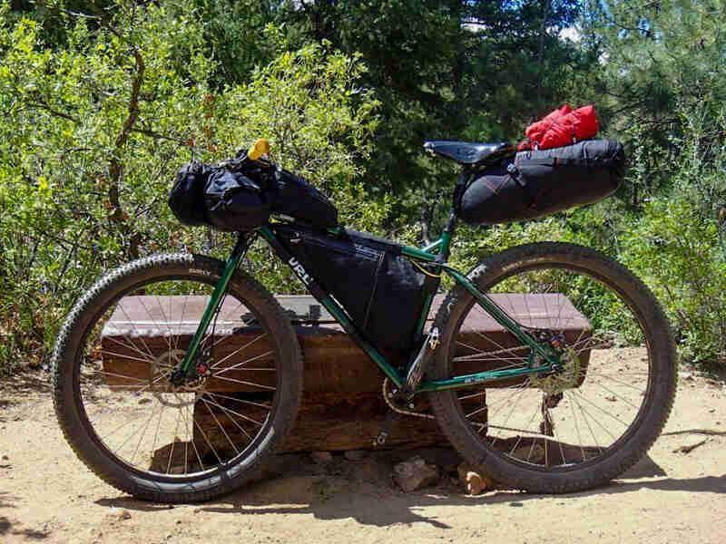 Left profile of a Surly Krampus bike, green, loaded with gear, in front of a bench, with the forest in the background