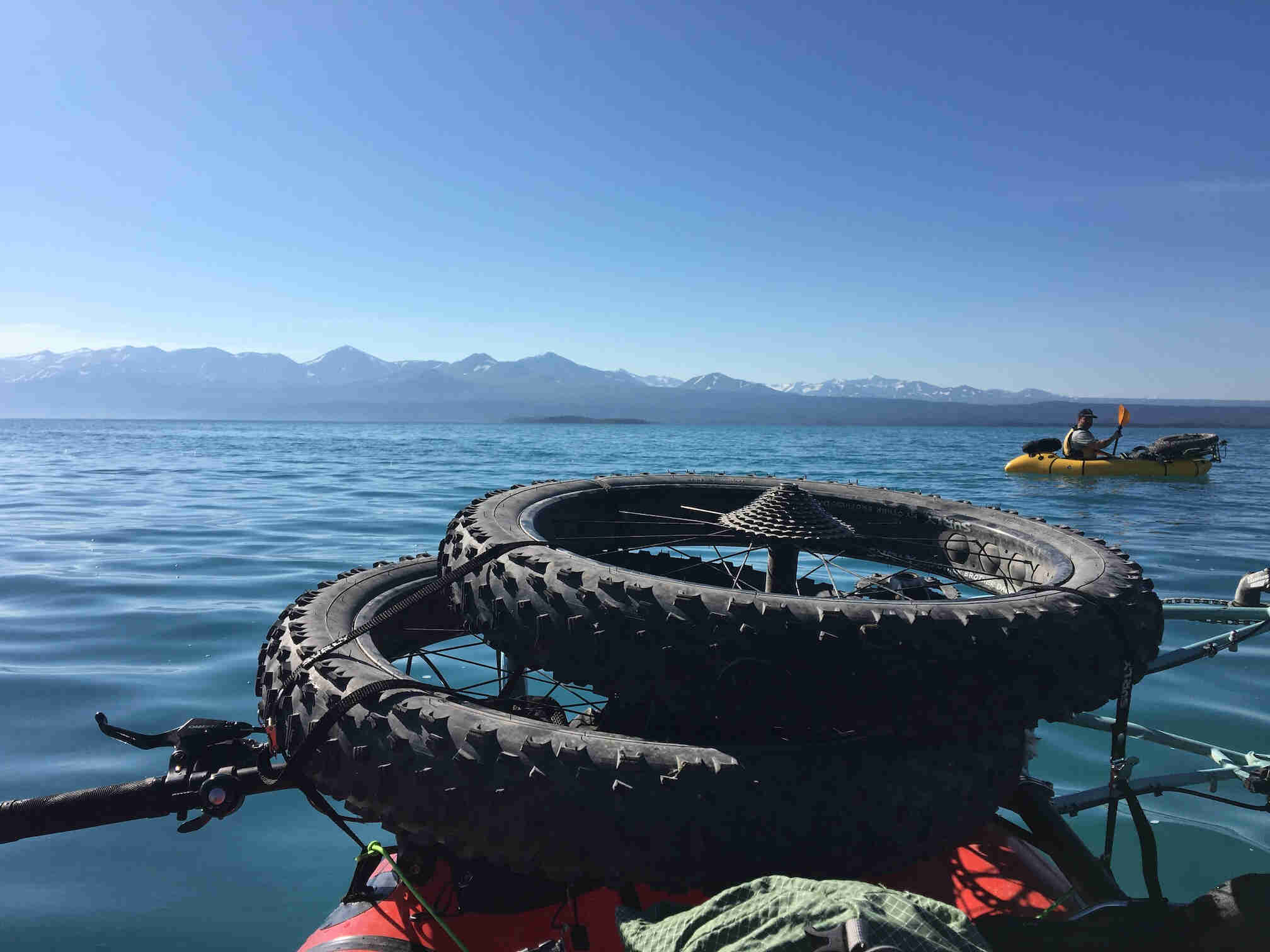 Close up of fat bike wheels on the front of a kayak on a lake, with mountains in the background