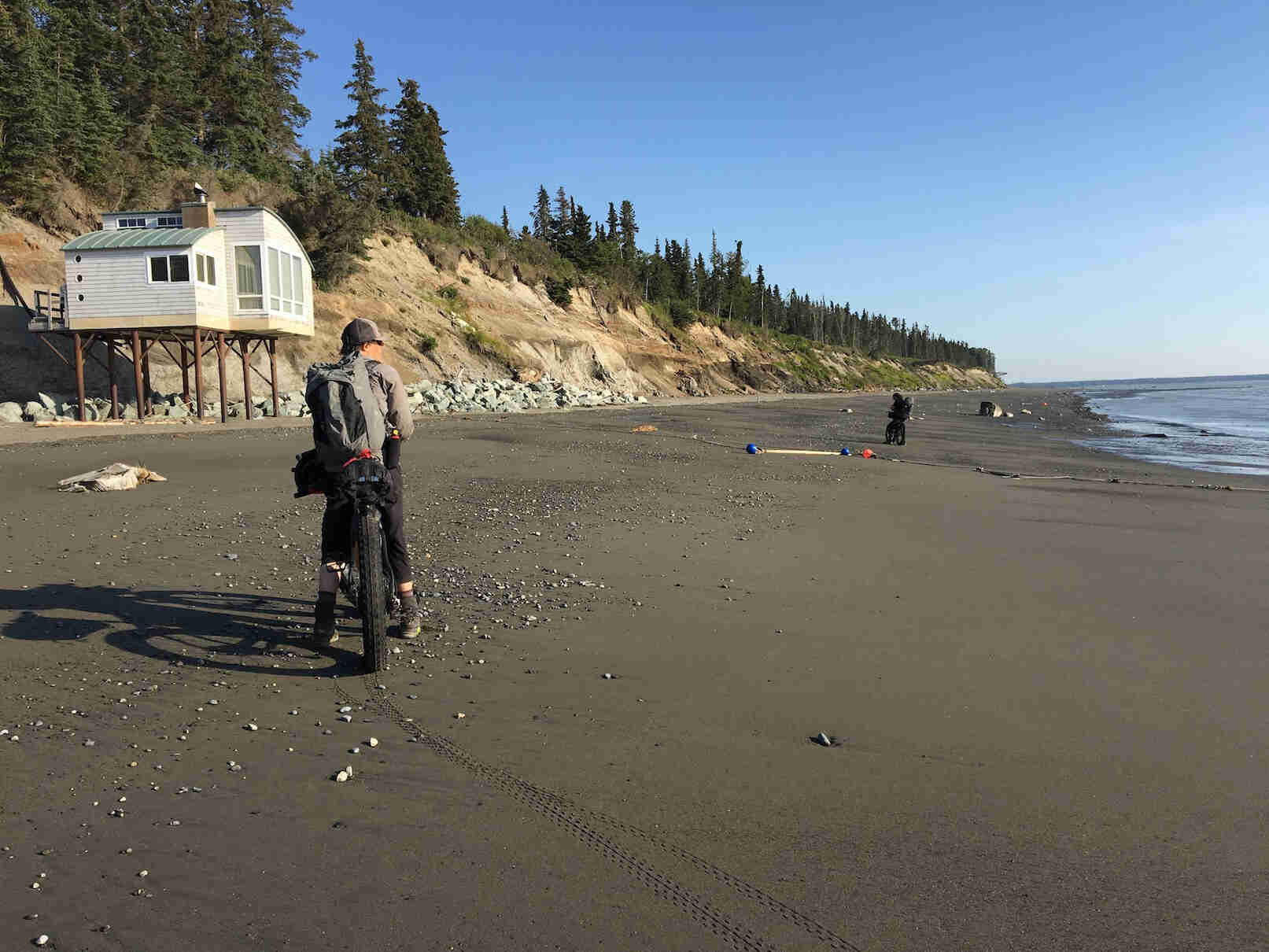 Rear view of a cyclist standing with their fat bike on a beach with water to the right, a building on stilts
