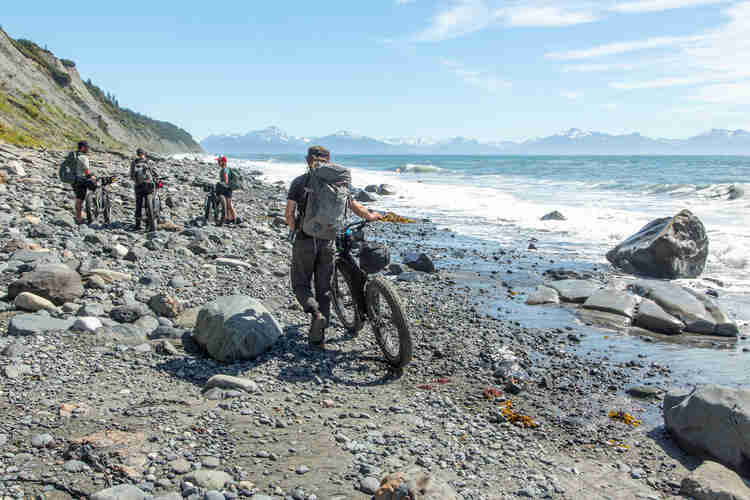 Rear view of a cyclist walking with a fat bike on a rocky shore towards 3 friends, with mountains in the background