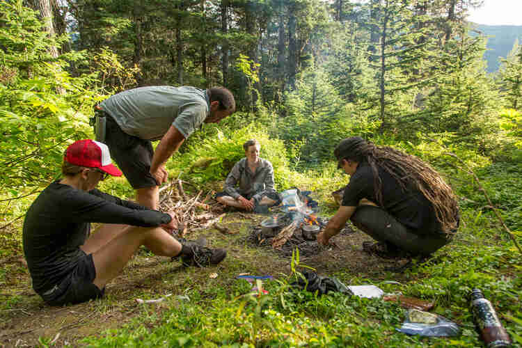 Four people sitting around a campfire on a grass clearing on a mountain hill, with trees in the background