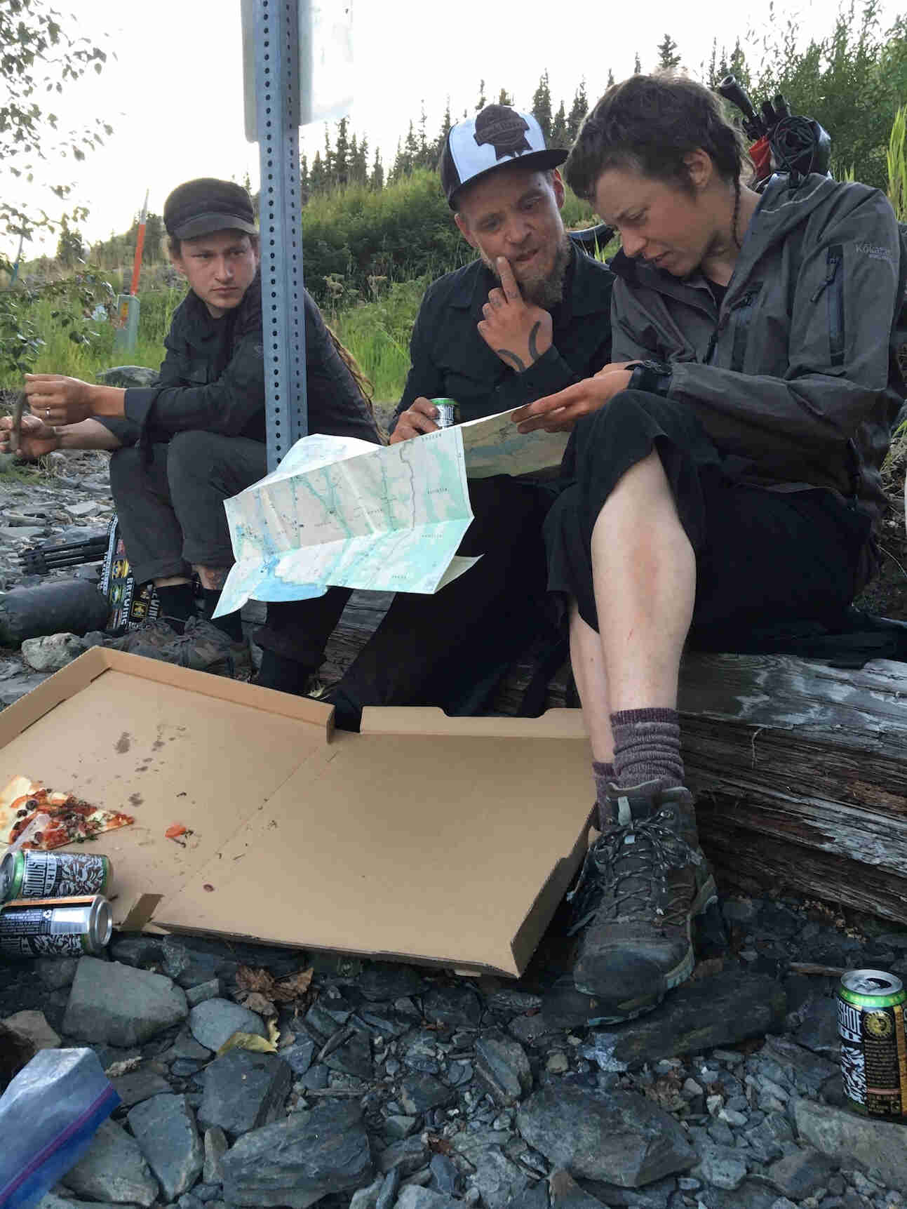 Front view of 3 people sitting on a log, looking at a map, with a pizza box sitting on the ground in front of them
