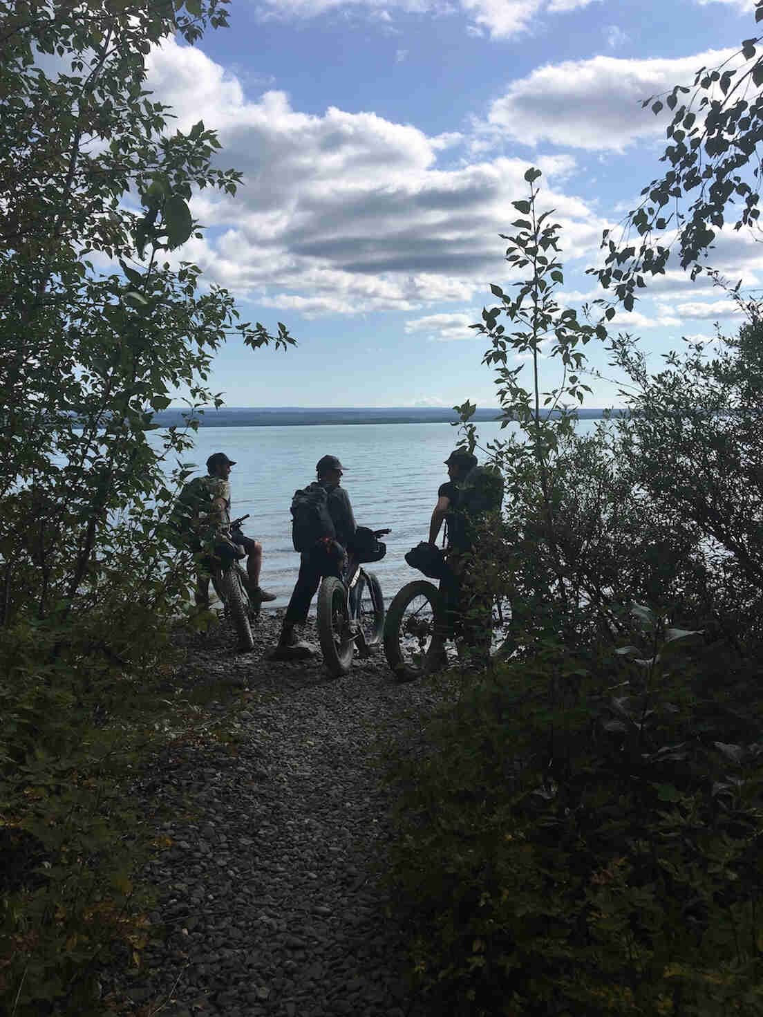 Rear view of three cyclists standing with their fat bikes, at the end of a gravel trail, in front of a lake