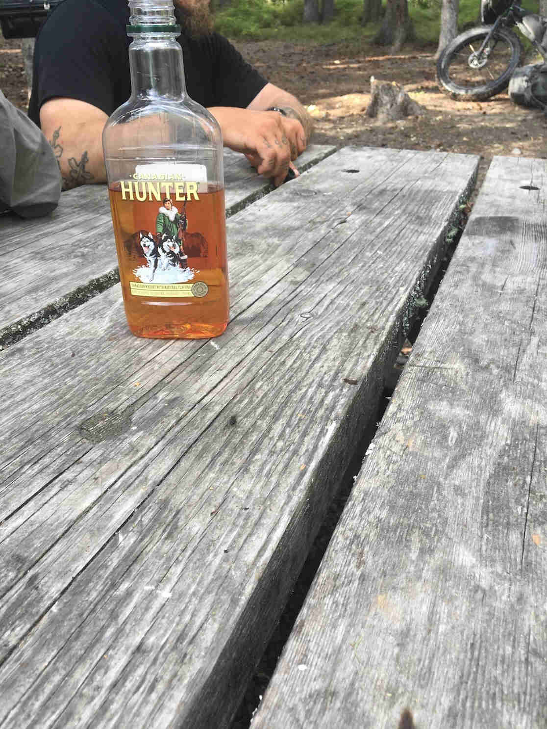 A bottle of Hunter liquors, on a picnic table, with a person sitting at the left end