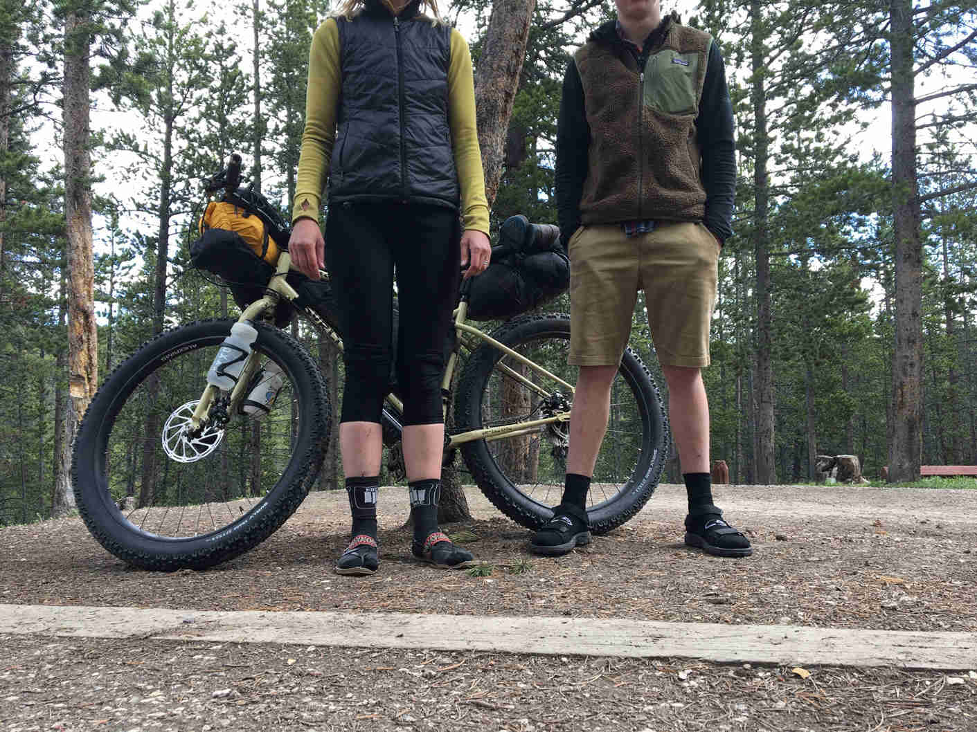 Two cyclists standing in front of a Surly ECR bike, on gravel, with pines trees in the background