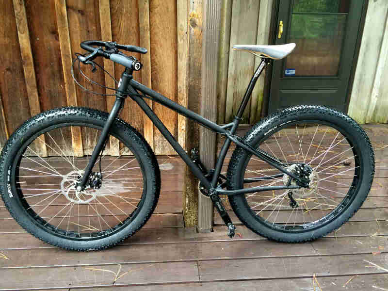 Left side view of a Surly Krampus bike, matte black, in front of a wood sided wall, on a porch