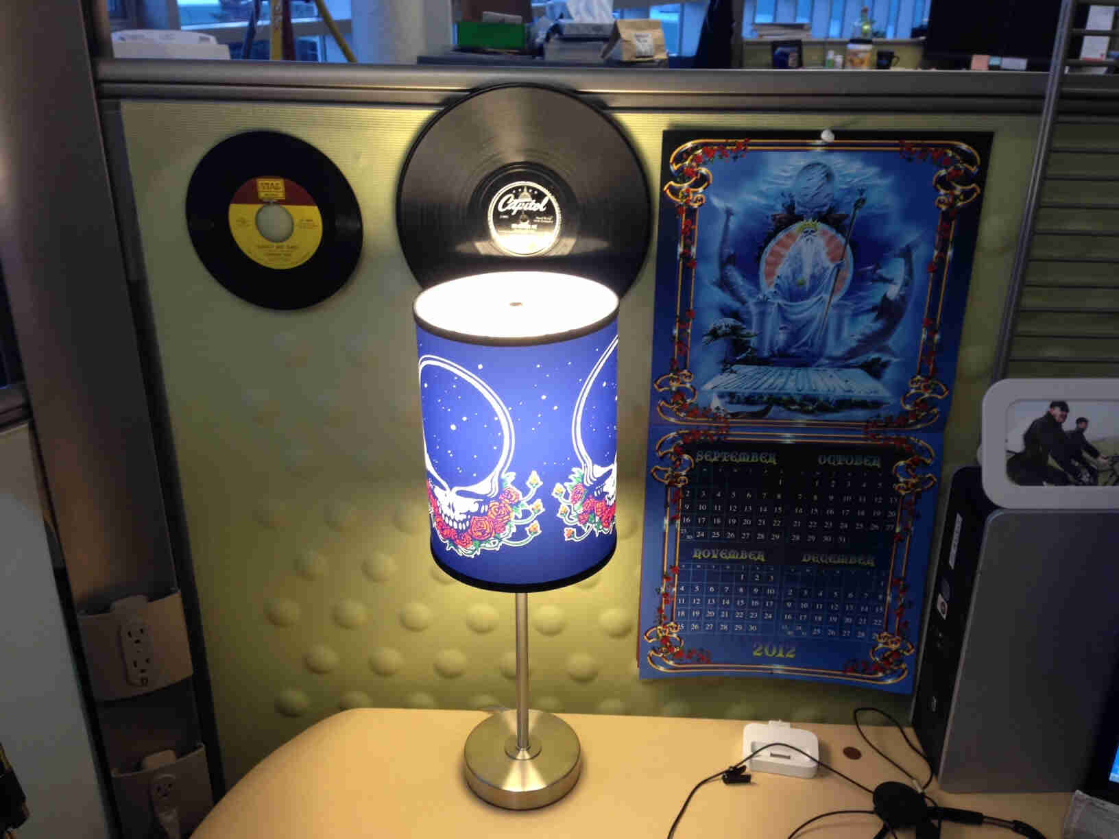 A lamp with a Grateful Dead shade, on a table in a cubicle, with records on a wall behind it