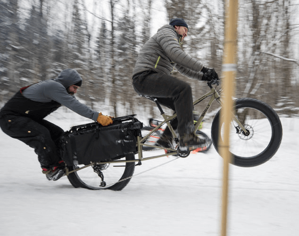 Right side view of a cyclist popping a wheelie on a Surly Big Fat Dummy bike in the snow, with a person hanging on back