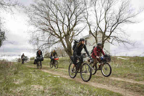 Front view of cyclists riding down an incline on a country dirt/grass road in front of a farmhouse