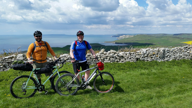 Two cyclist standing behind their bikes, right side view, with a stone wall and the sea in the background