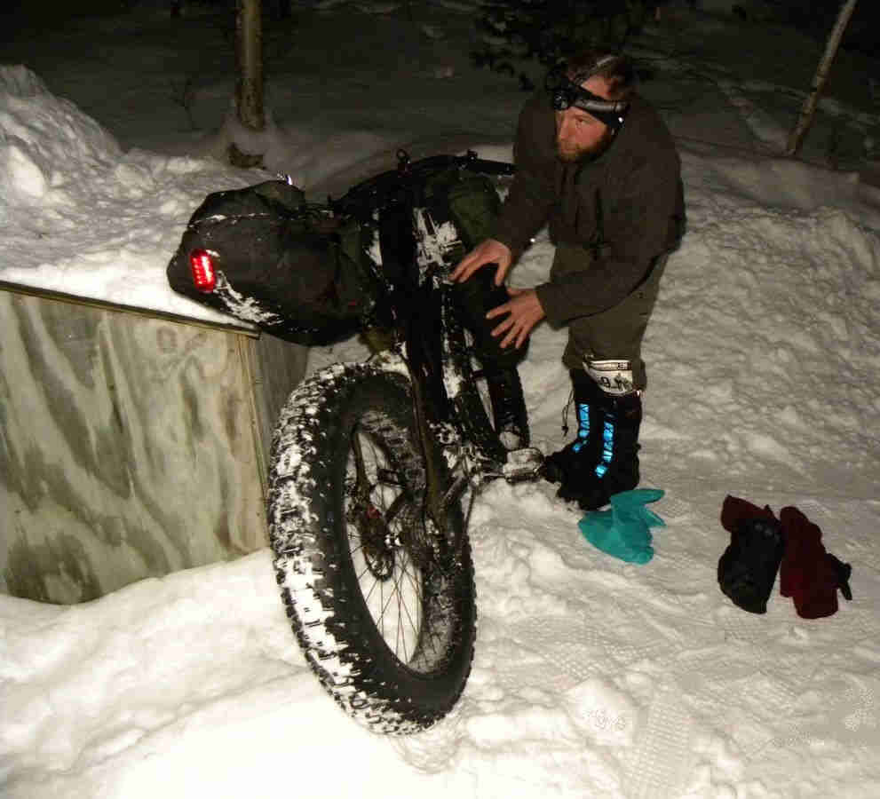 Rear, right side view of a Surly Moonlander bike, on deep snow, with person loading gear on the right side at night