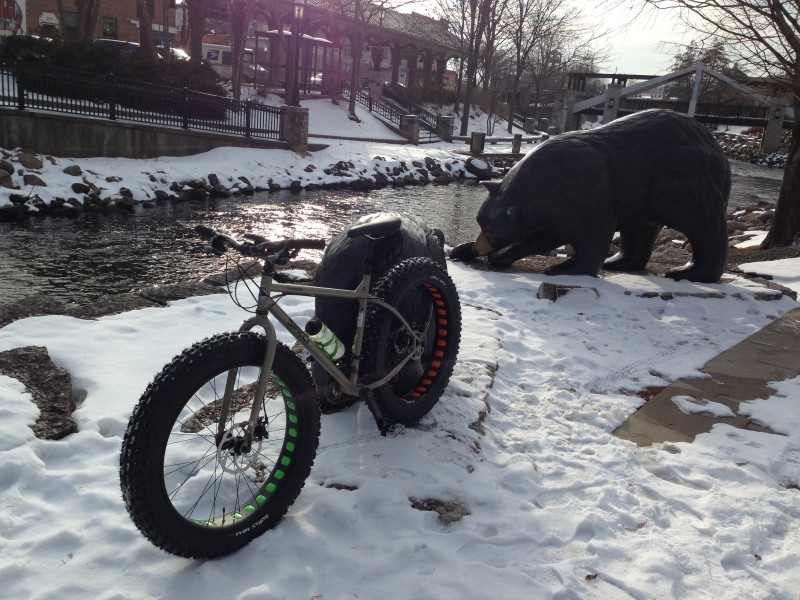 Left side view of a Surly Moonlander fat bike, parked in snow next to a stream, with a brown bear statue in background