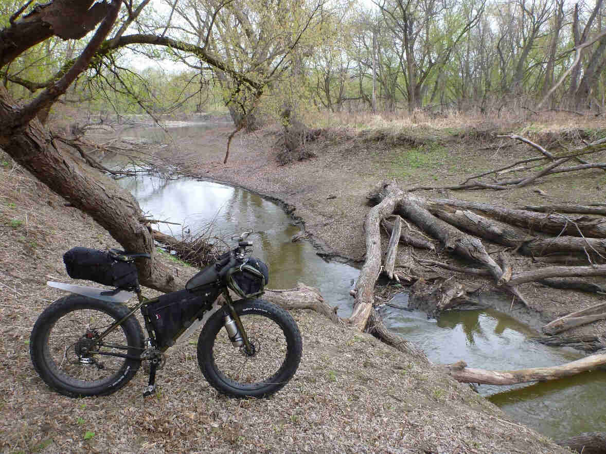Right side view of an olive drab Surly Moonlander bike, parked on a bare bank, facing a stream with a downed tree in it