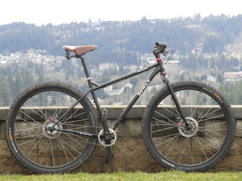 Right side view of a black Surly Karate Monkey bike, parked along a stone block wall, with a town on a hill behind it