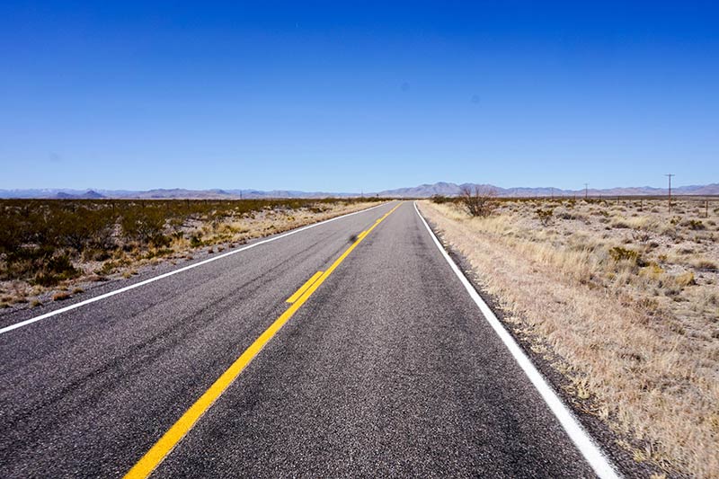 A straight, paved two lane highway in the desert heads into the mountain with blue sky above