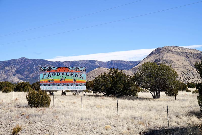 A rainbow colored Magdalena Billboard stands in prairie grass field with trees and mountains in the background