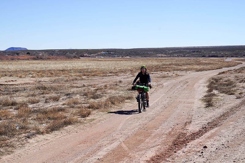 A front view of a cyclist riding on a gravel road in a grassy desert on a clear day