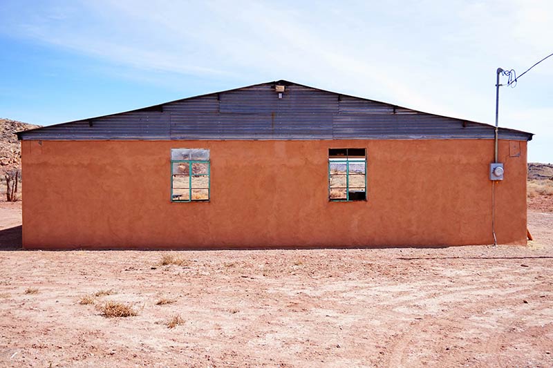 An orange stucco building with a steel roof sits on a sand lot in the desert with blue sky above
