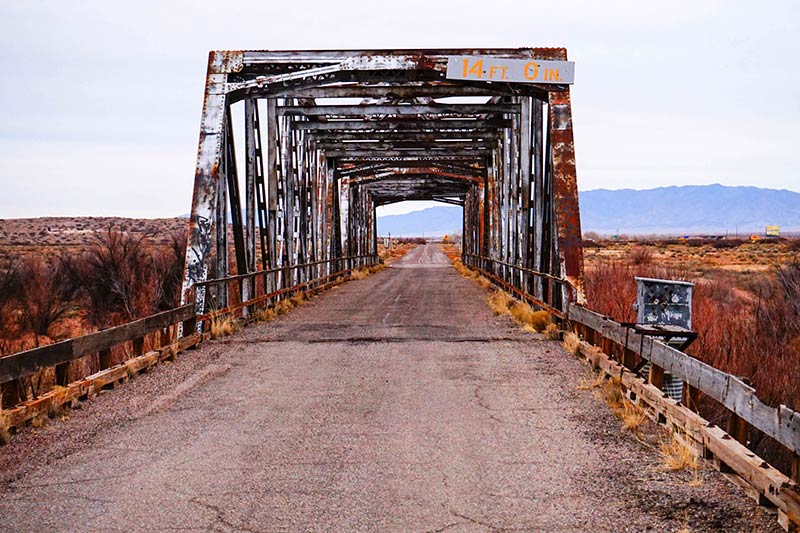 A straight view of a rusty steel bridge,  with a road running through and brush on each side, and mountains ahead