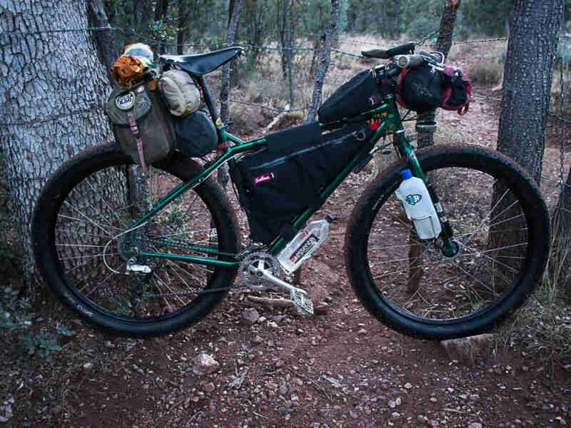 Right side view of a green Surly Krampus bike with gear, parked across a dirt trail in the forest, between 2 trees