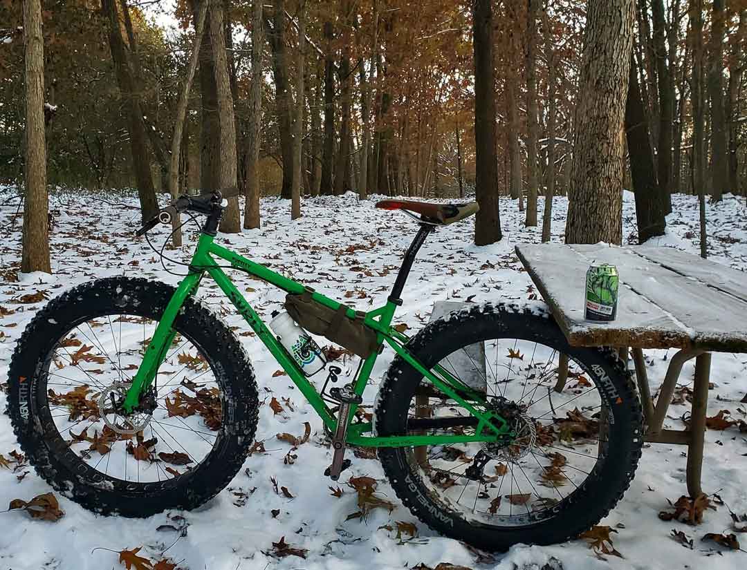 Left side view of a green Surly Pugsley fat bike, with the back tire wedged under a picnic table, in snowy woods