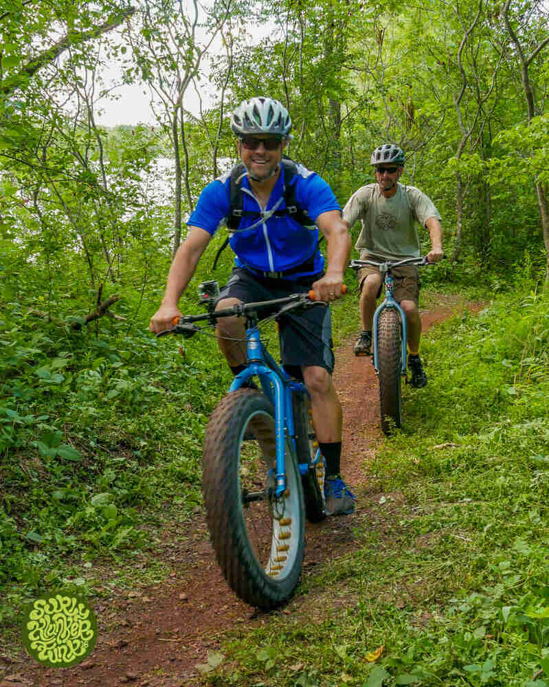 Front view of 2 cyclists riding Surly fat bikes, on a dirt trail in the woods