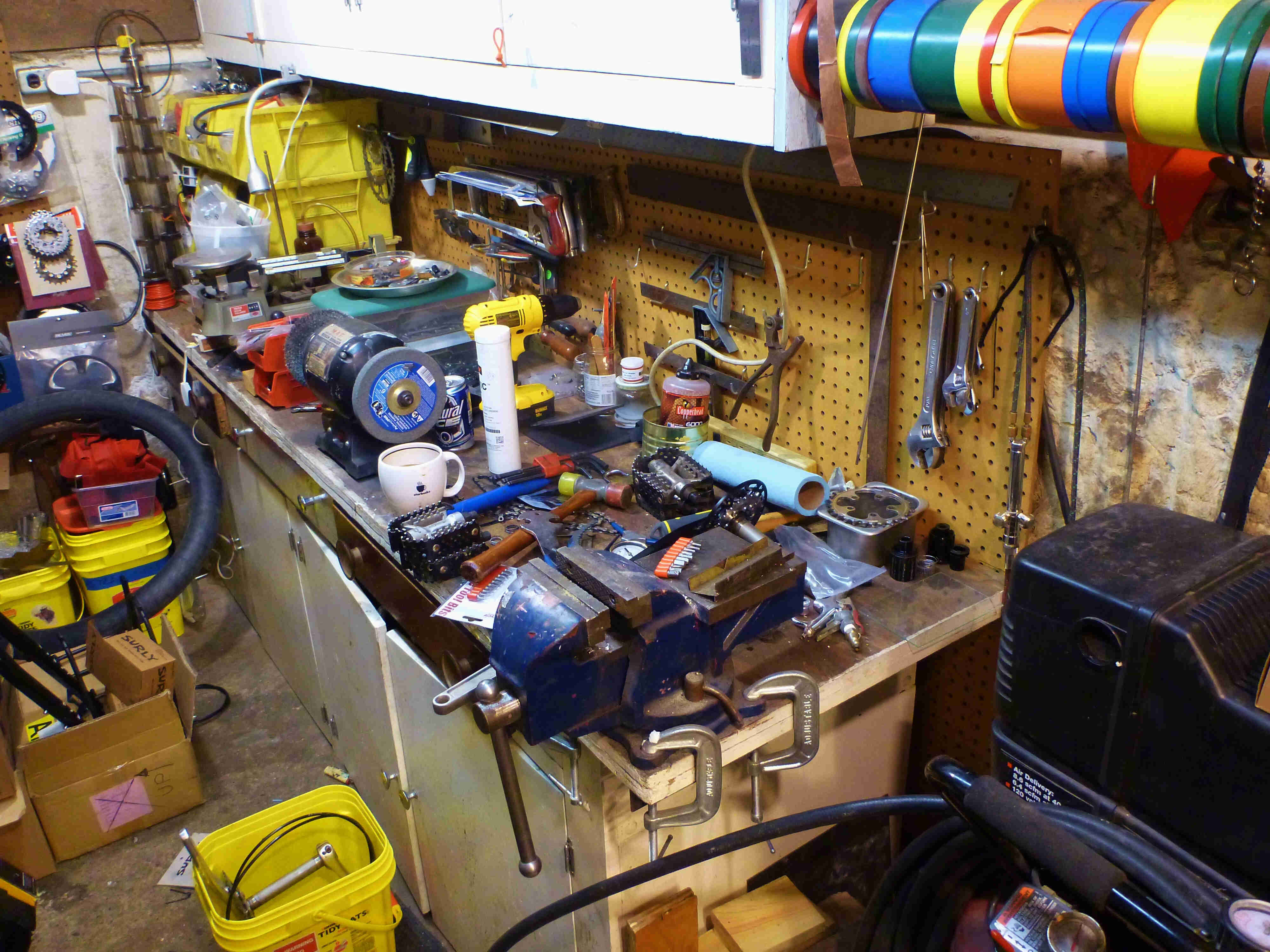 A workbench, in a workshop, with a vise, a grinder mounted to it, and bike parts laying all around