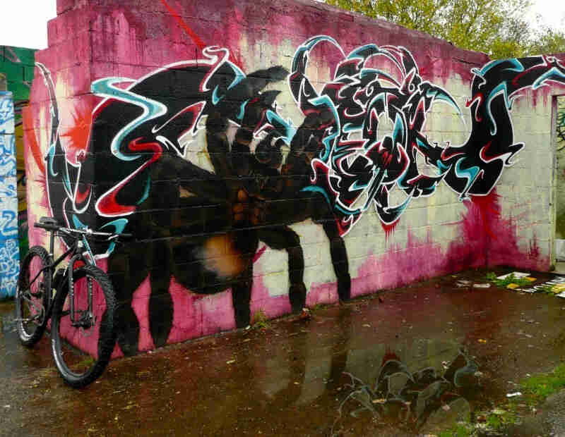 Front, right side view of a Surly bike, parked on corner of cinder block walls, with graffiti painted on the front wall