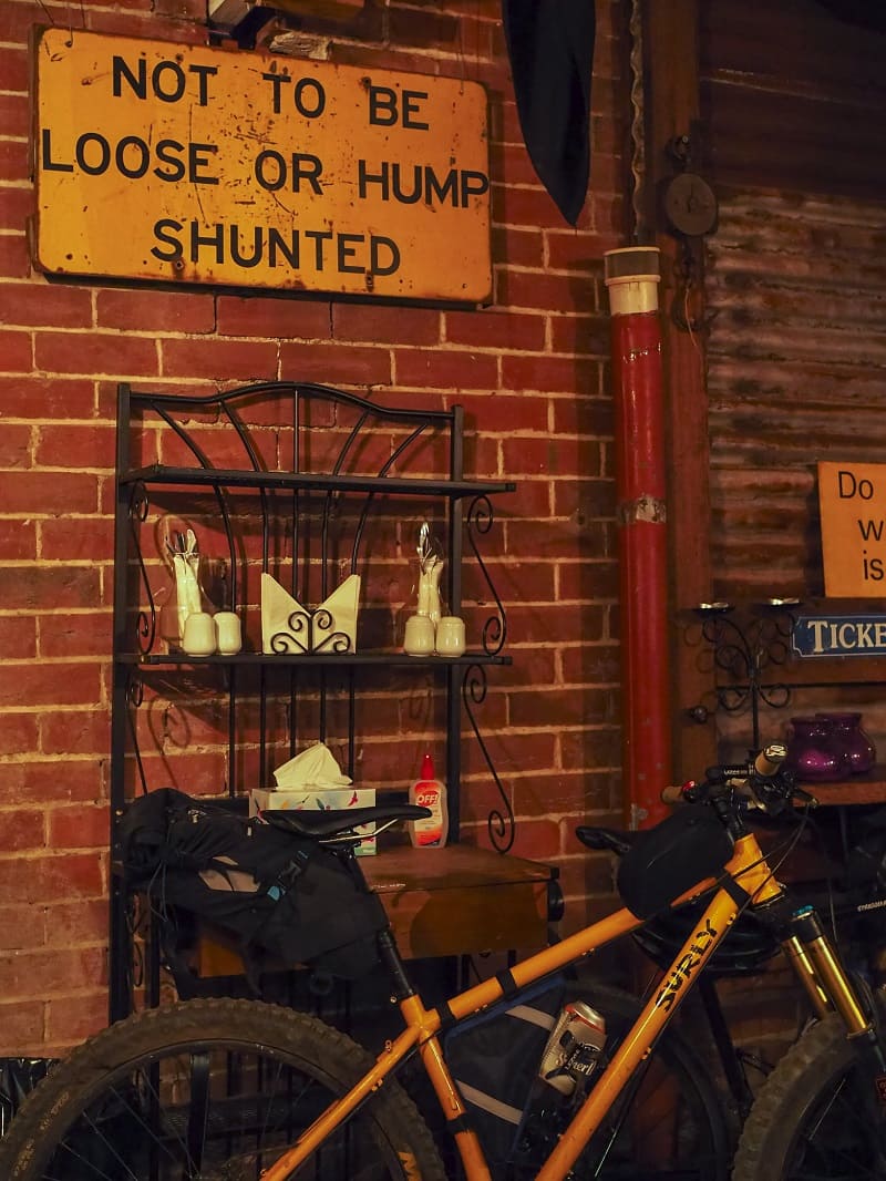 Right side view of a yellow Surly bike in front of bakers rack, sign against a brick wall