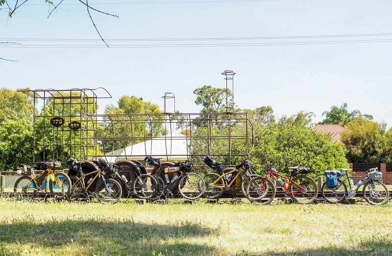 Right side view of bikes in a single file line, with a railroad track and steel tube train sculpture, with trees behind