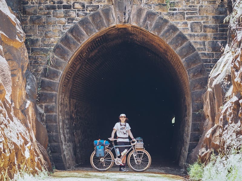 Cyclist standing behind a bike loaded with gear, shown in a right profile, at the entry to a mountain tunnel