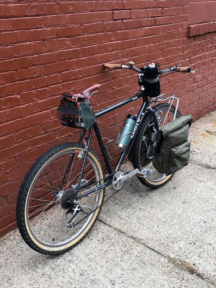 Right side view of Surly Pack Rat bike loaded with gear, leaning against a red brick wall on a sidewalk
