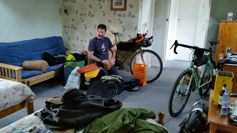 A person sitting in a chair in a room with gear scattered around and 2 bikes