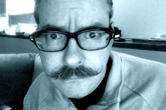 Headshot of a person with a twisted up mustache and wearing glasses - black & white image
