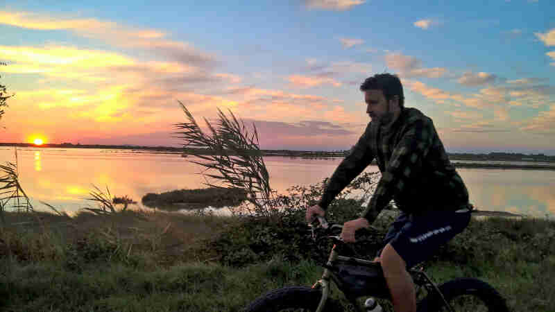 Left side view of a cyclist riding on a flat, grassy river bank, with the sun on the horizon in the background