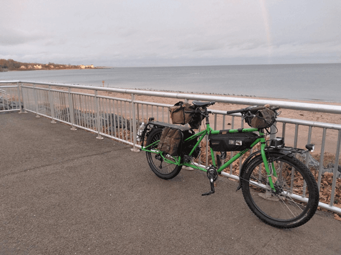 Left side view of a green Surly Big Dummy bike with fenders loaded with gear bags, leaning on guardrail above a beach