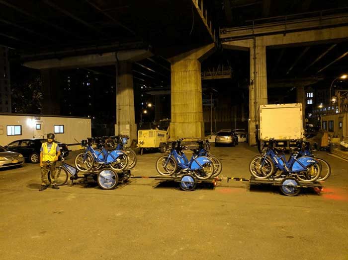 Person standing under a bridge at night in front of a three wheeled bike with two trailers loaded with bikes connected