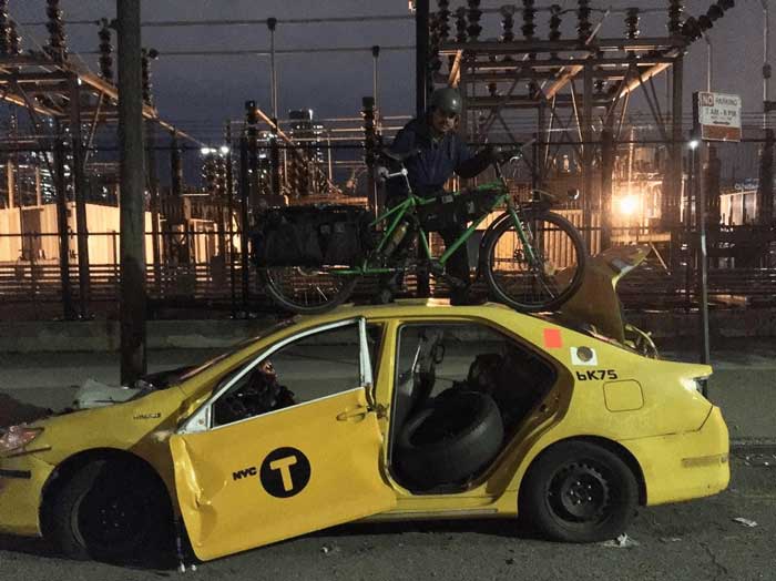 Cyclist with a green Big Dummy bike on the roof of a yellow broke down NYC taxi at night in front of an electric station