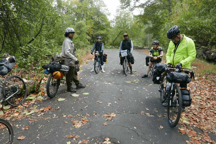 Four cyclists standing with their bikes loaded with gear on a paved trail in the woods