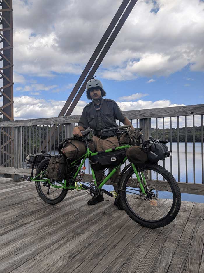 Cyclist standing on a wood bridge over a river with a green Surly Big Dummy bike with blue sky and clouds in the sky