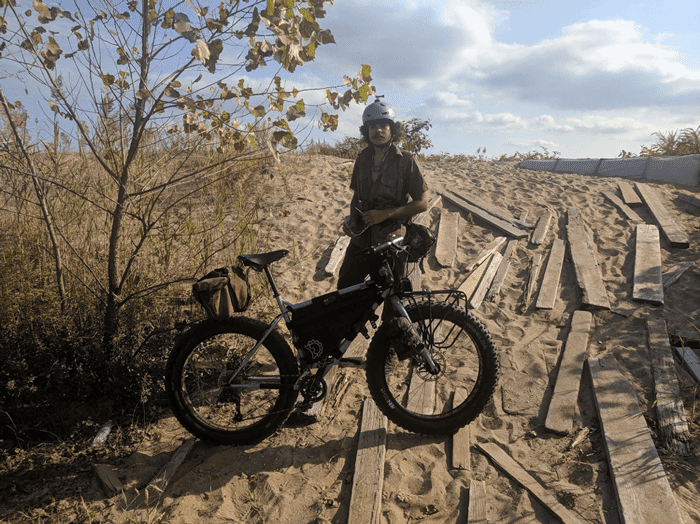 Right profile of a white Surly Pugsley fat bike loaded with gear and cyclist standing behind on a sandy plot with boards