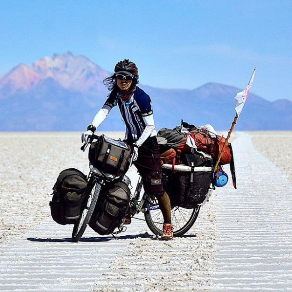 Cyclist facing toward camera while standing on a sandy road with a bike loaded with gear and mountains behind