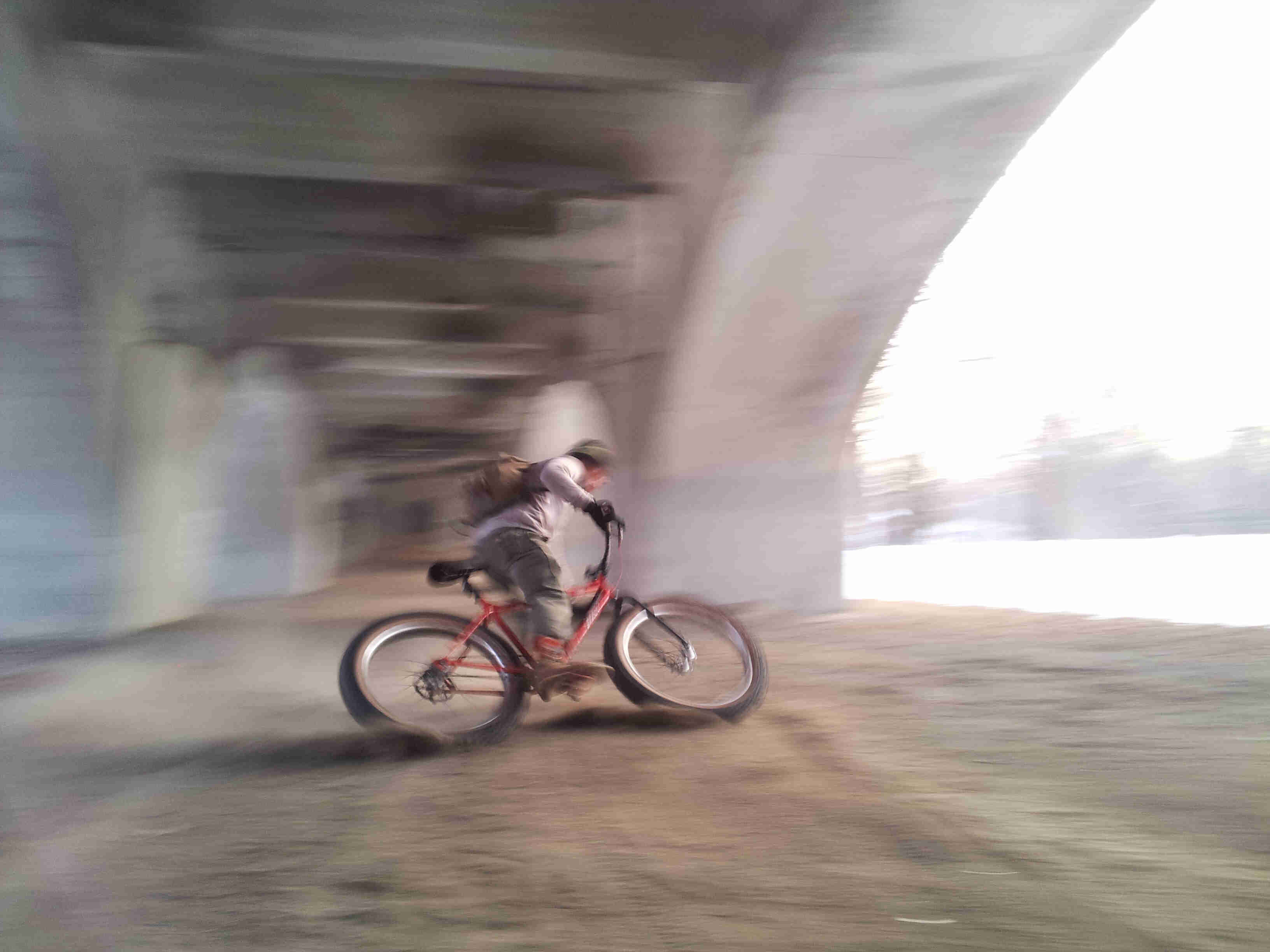 Blurry, right side view of a cyclist, leaning into a left turn on their bike, underneath a bridge overpass