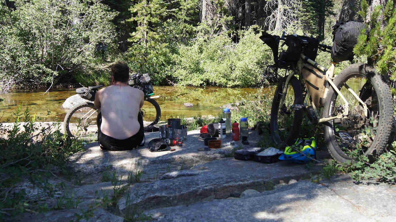 Rear view of a person sitting on a flat rock, facing a stream, with a Surly fat bike in front of them and another behind