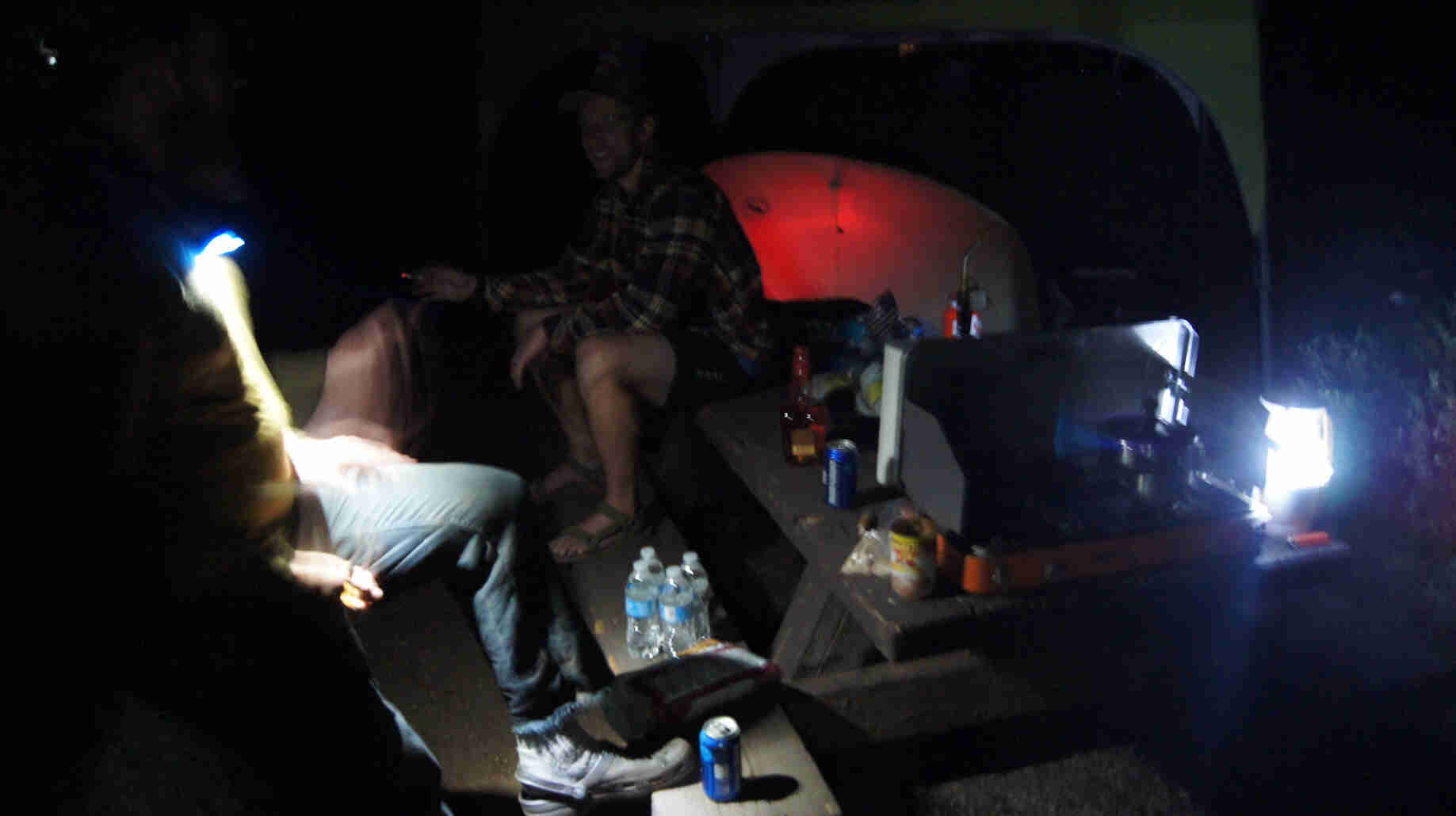 Two people sitting on a picnic table with a tent in the background, in a campsite at night