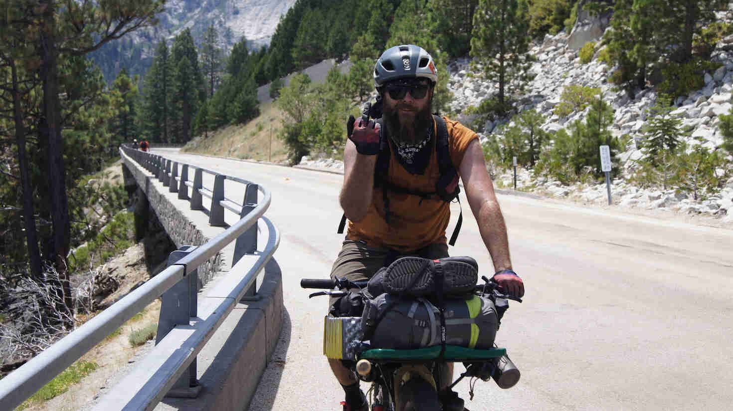 Front view of a cyclist, holding up their middle finger, riding up a hill on a paved, with tree covered mountains behind